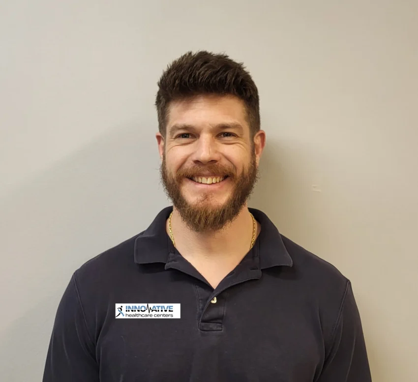 Smiling man in a dark polo standing against a neutral wall, wearing a logo patch for Innovative Healthcare Centers on his chest.