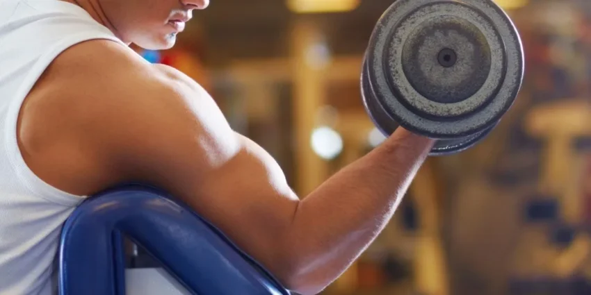Why Bicep Curls Can Irritate the Inner Elbow - Innovative Health Care Centers Person in a white sleeveless shirt performing a bicep curl with a dumbbell at the gym, seated on a bench.