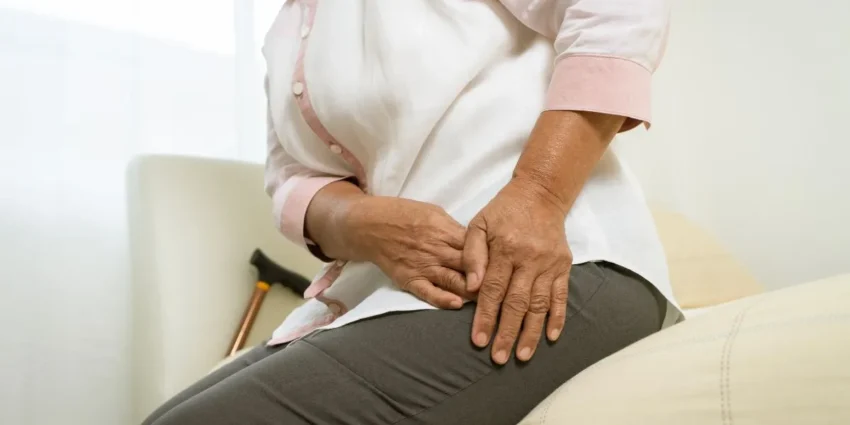 Person seated on a bed, holding their knee with both hands as if in pain or relief.